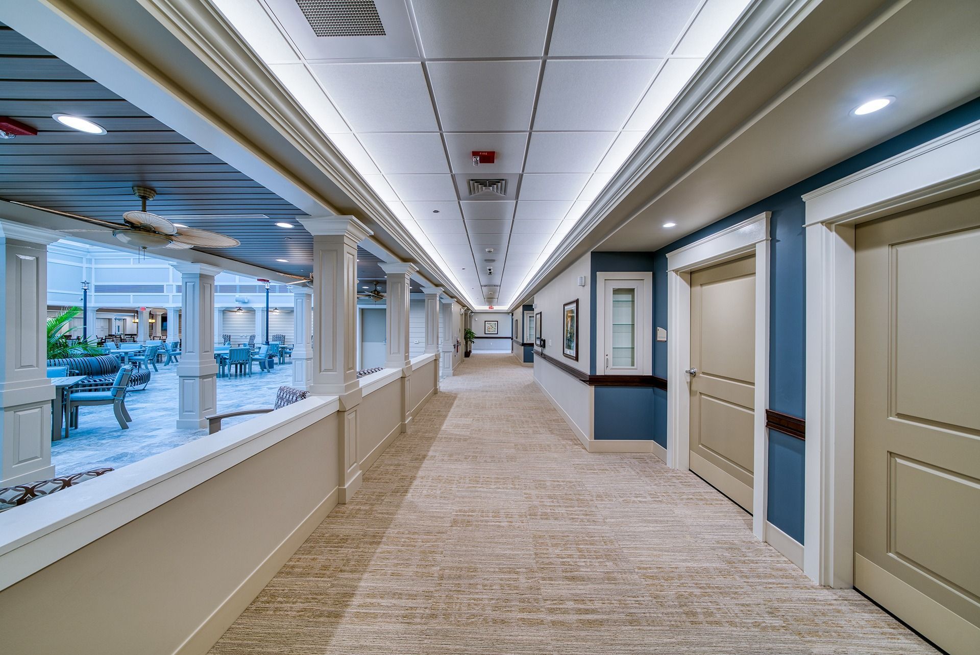 Bright hotel corridor with beige carpet, white walls, blue accents, and windows overlooking a pool.
