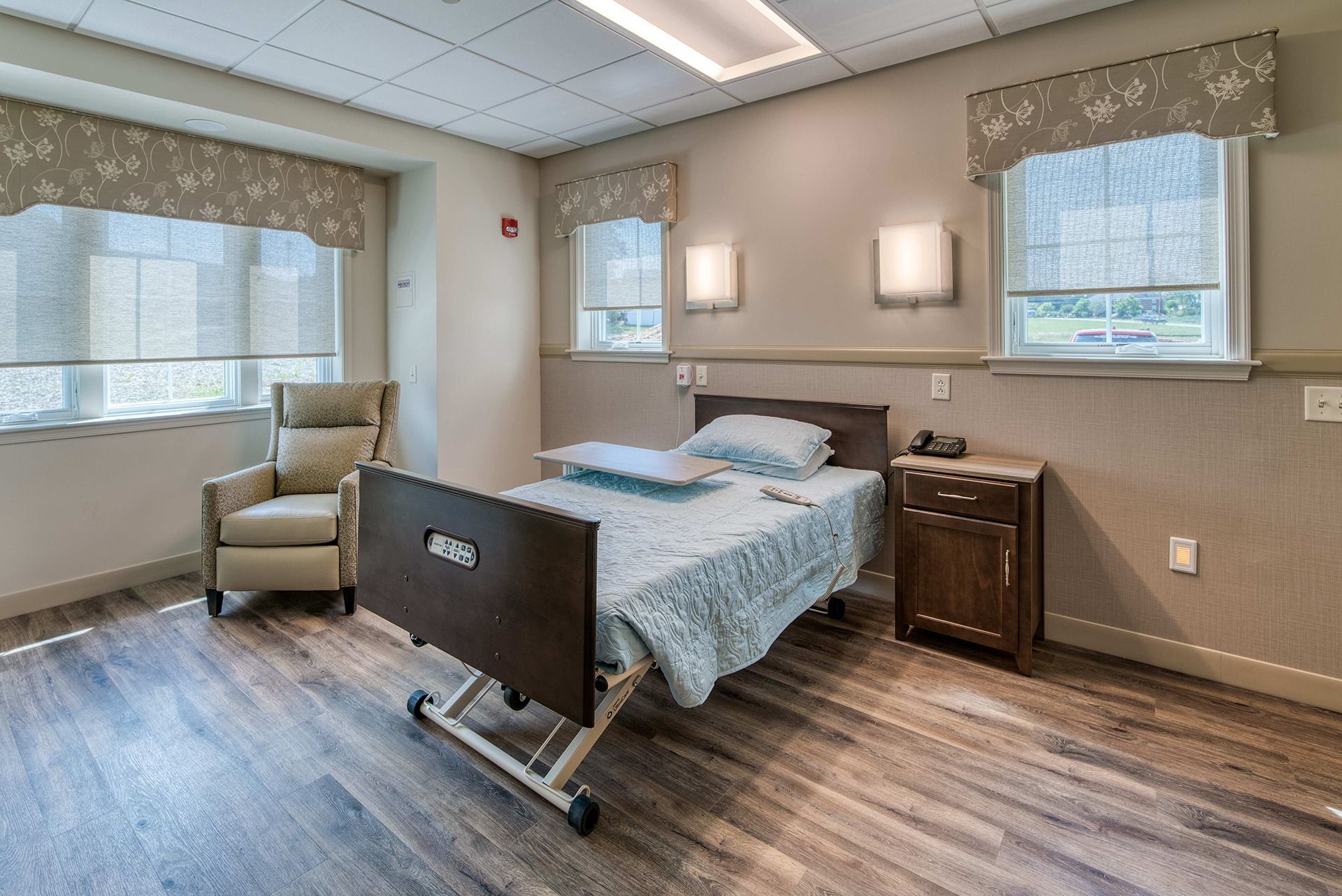 Empty hospital room with bed, recliner, nightstand, and large windows.