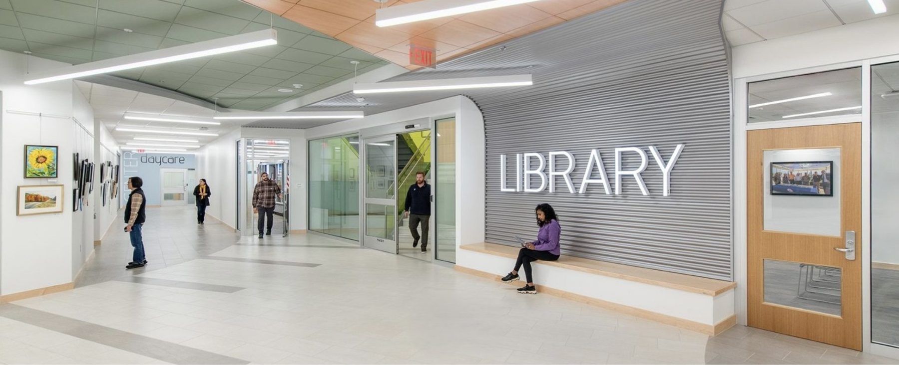 Modern library hallway with glass walls, large “LIBRARY” sign, and people walking and sitting nearby