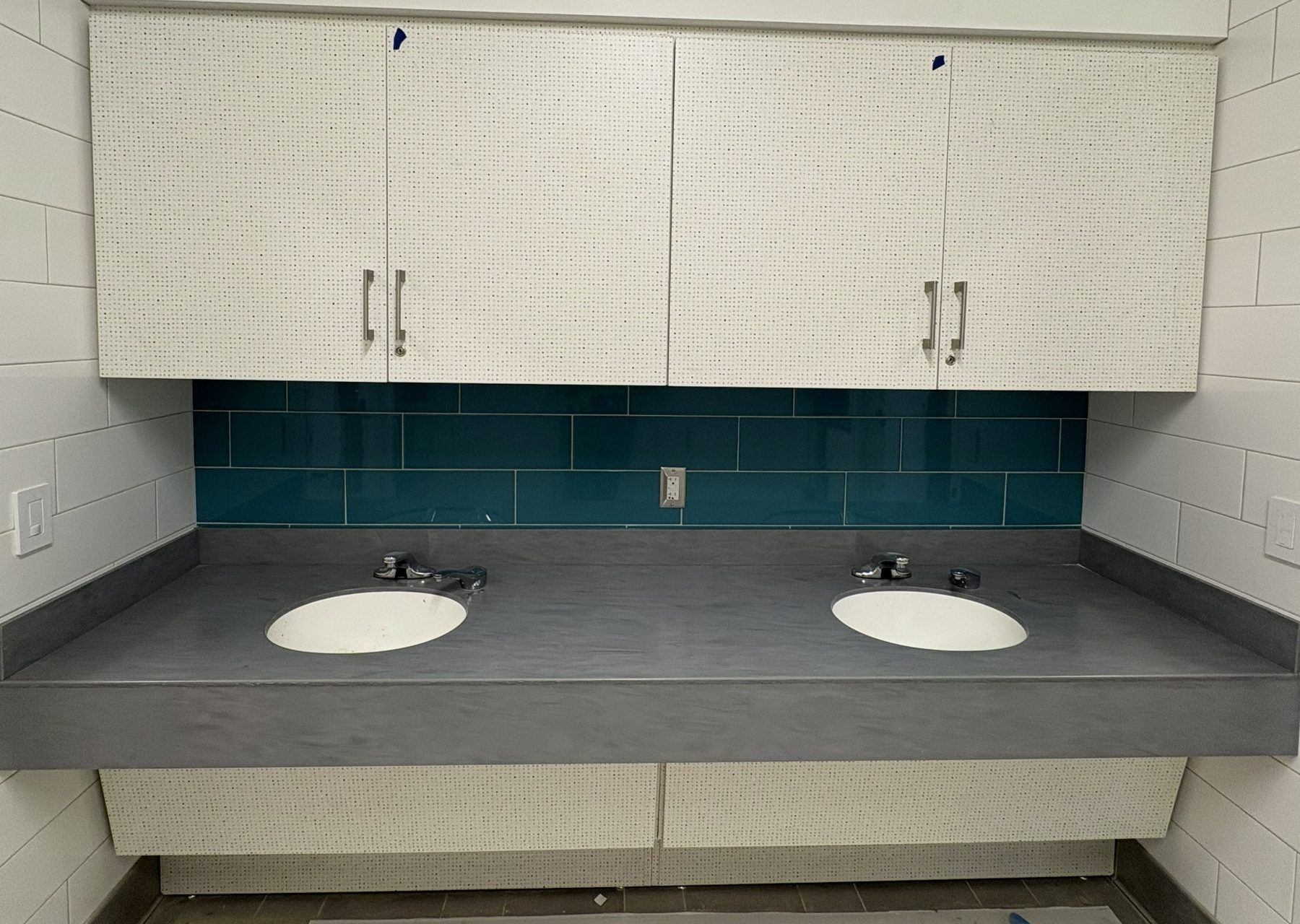 Bathroom vanity with two sinks, gray countertop, teal tile backsplash, and white cabinets above and below.