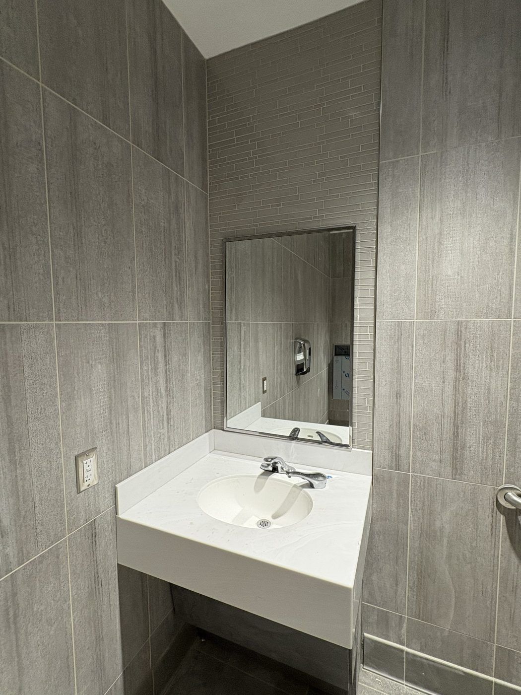 Small restroom sink with mirror, gray tiled walls, and white countertop.