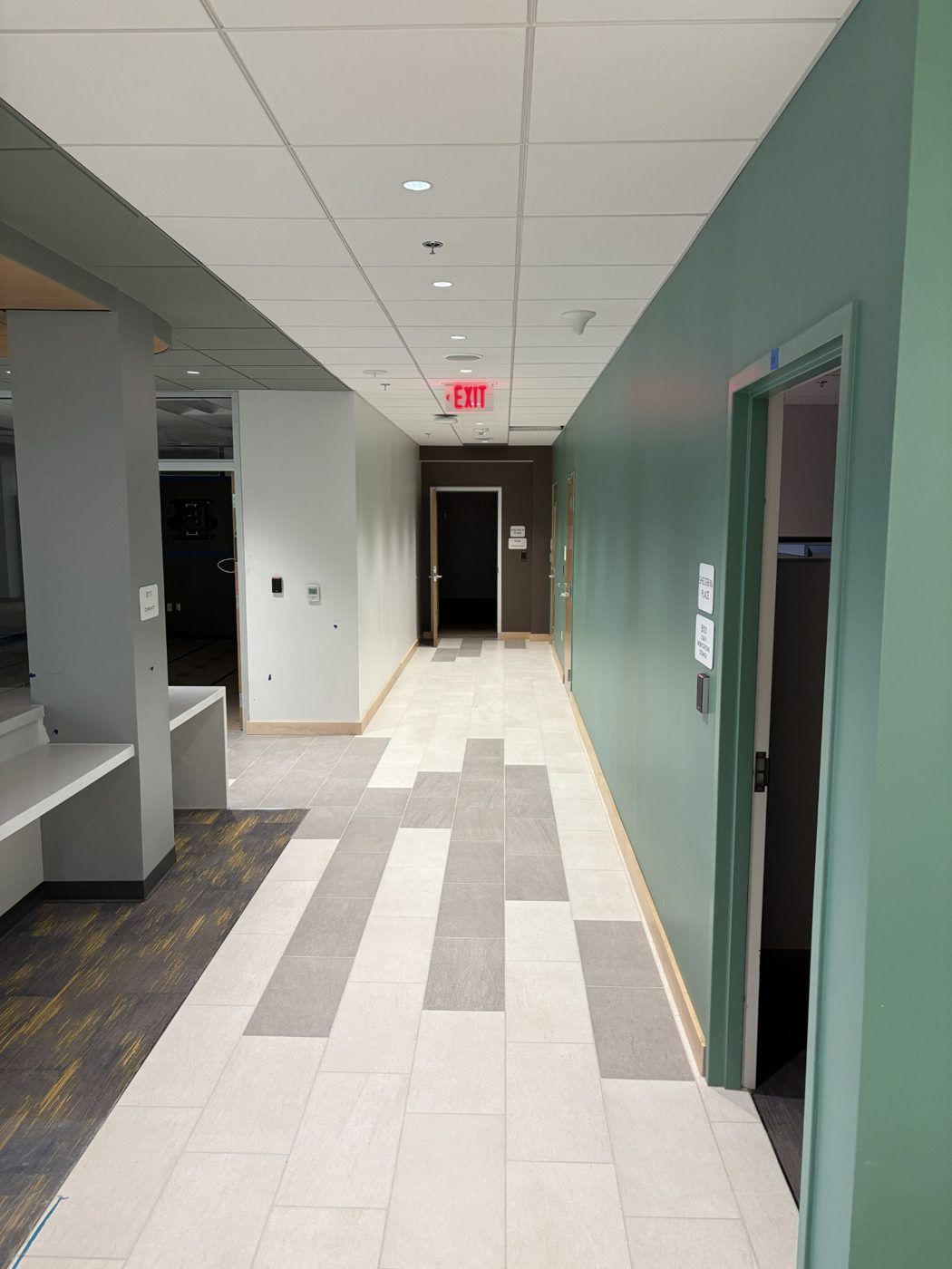 Empty hallway with tiled floor, teal walls, overhead lights, and a red EXIT sign at the far end