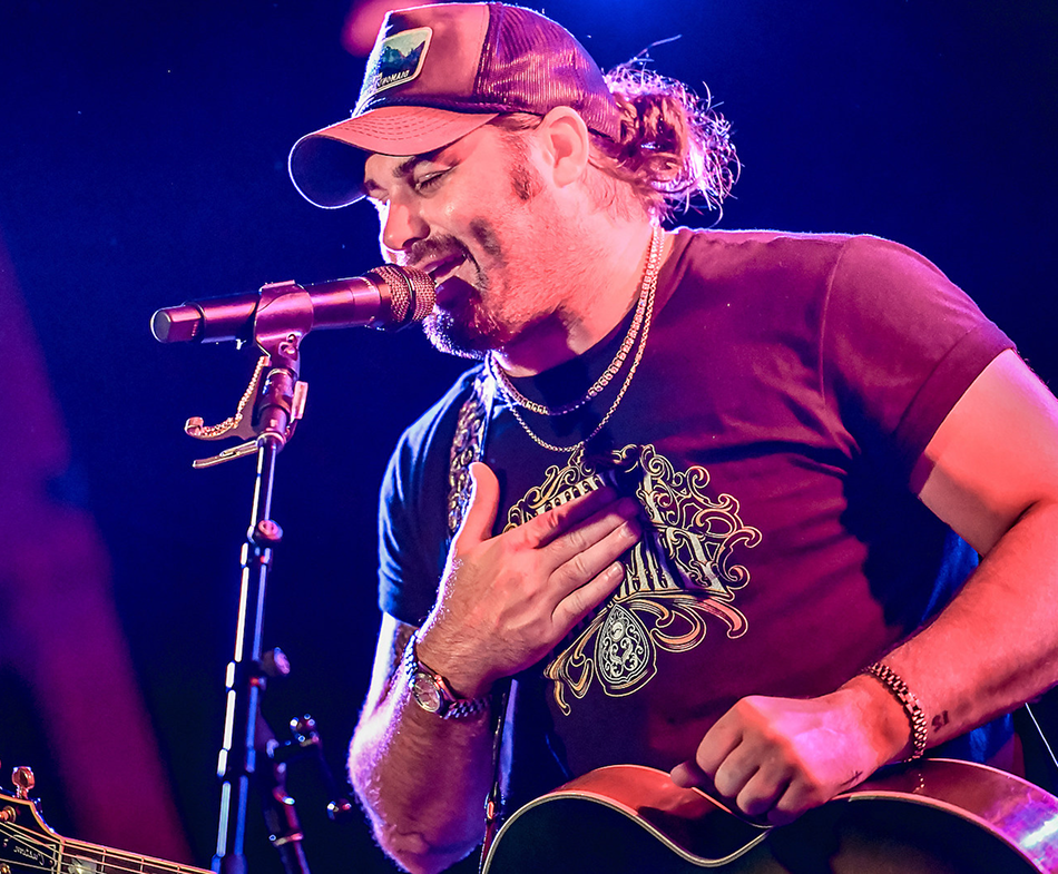 Singer in a cap performing into a microphone under purple stage lights, hand on chest, holding a guitar.