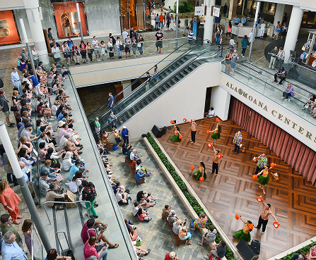 Crowded indoor mall atrium with people on escalators, balconies, and a performance area below