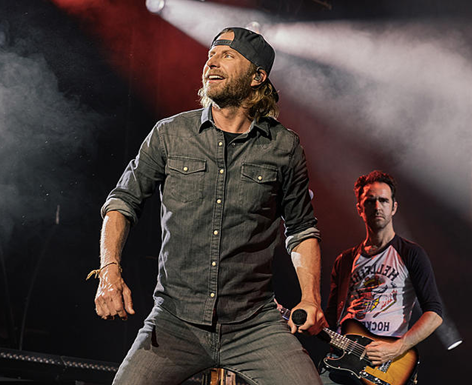 Singer on stage under bright lights, wearing a dark shirt and cap, with a guitarist in the background.