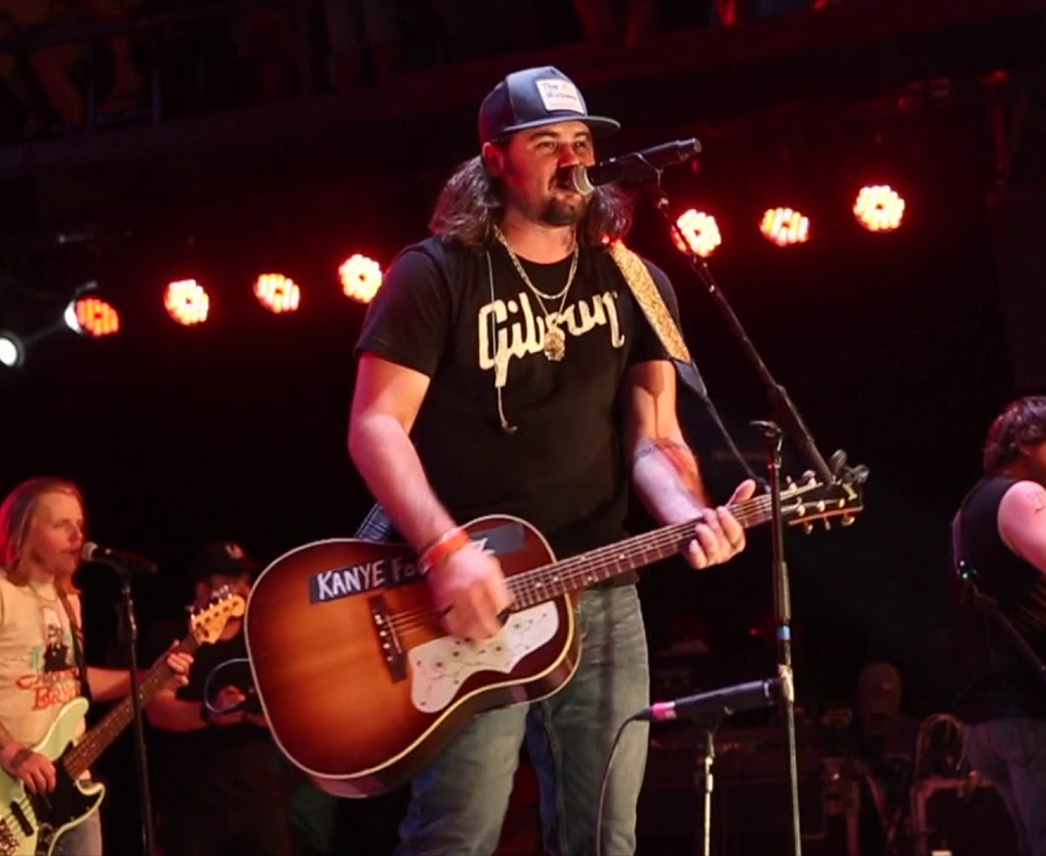 Singer-guitarist performing on stage under red lights, wearing a black Gibson shirt and cap, at a microphone