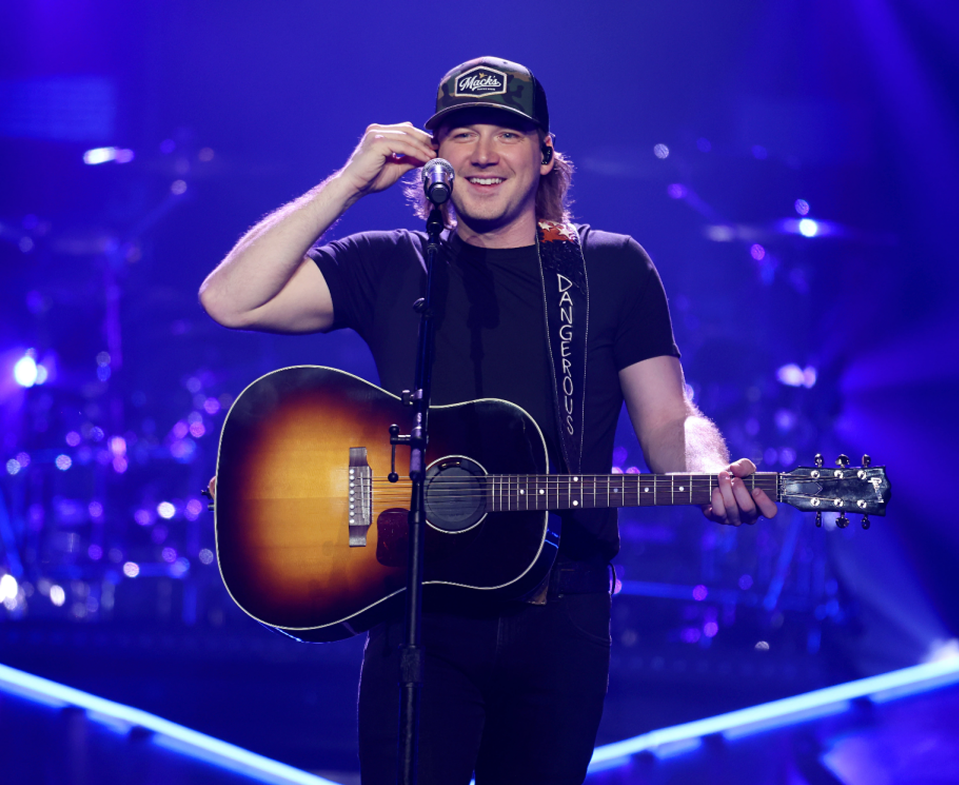 Singer holding a guitar on a purple-lit stage, smiling under a black cap.