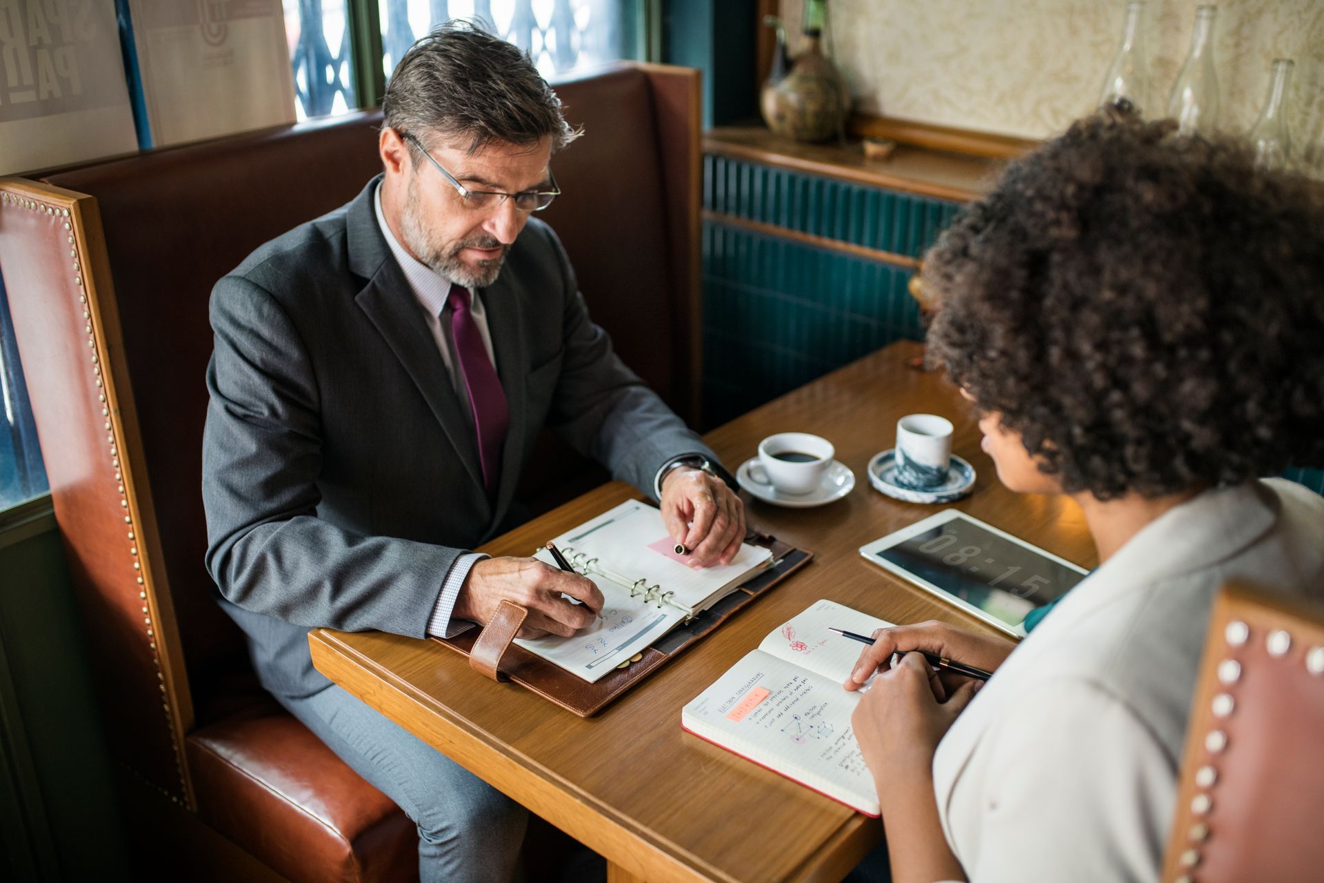 Two people discuss documents at a café table with coffee cups and a tablet.