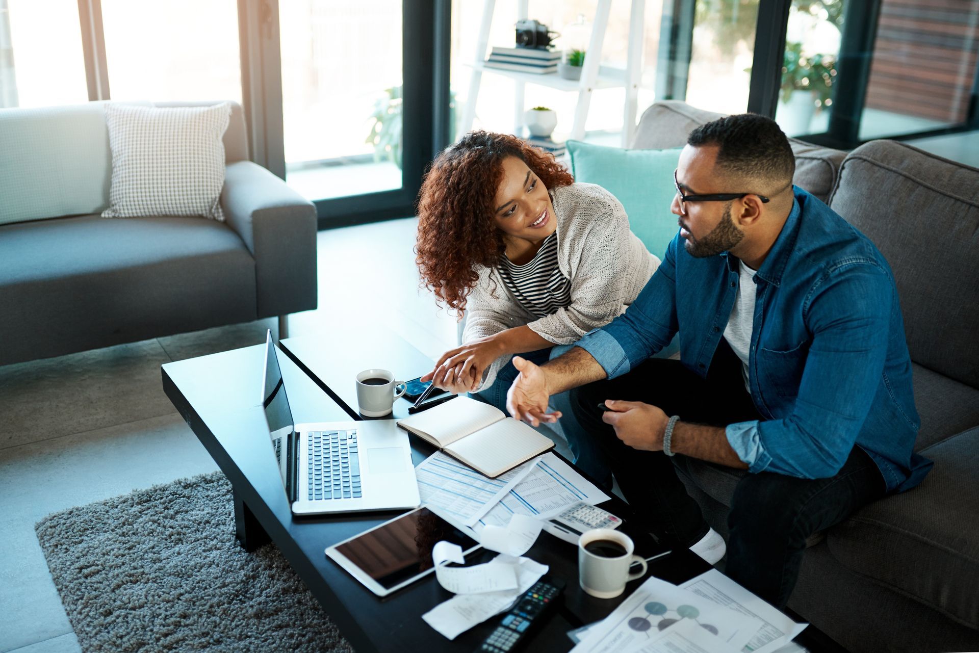 Two coworkers discuss papers beside a laptop on a living room coffee table.