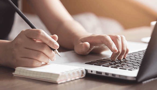 A person is writing in a notebook while typing on a laptop computer.