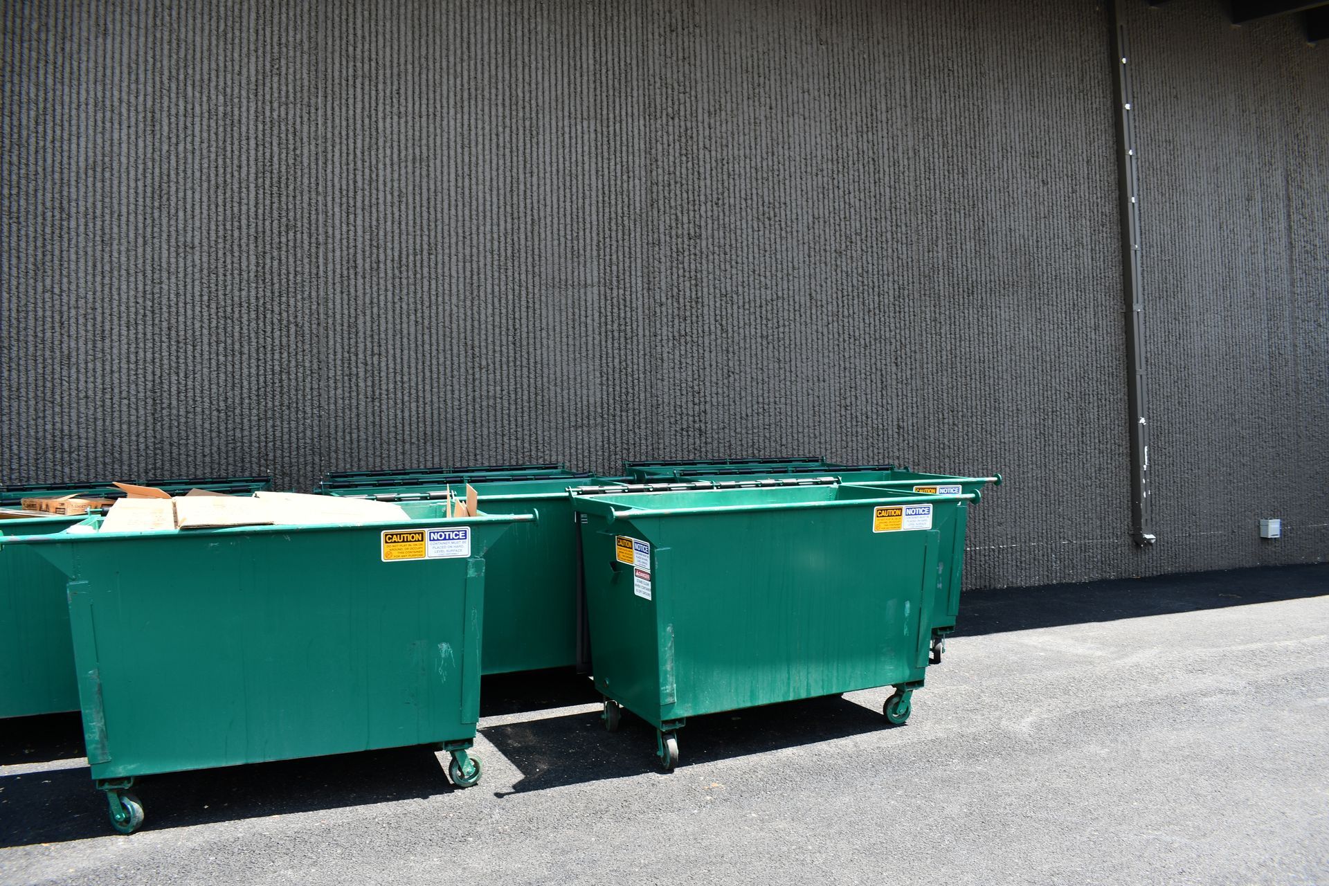 Green commercial recycling dumpsters with caution labels lined up against a gray building wall Green commercial recycling dumpsters with caution labels lined up against a gray building wall