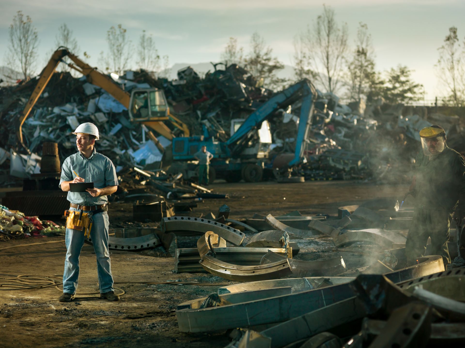 Worker in a hard hat logging scrap metal piles at a recycling center as excavators sort behind him