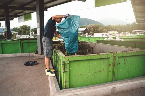 Man dumping yard waste into green recycling container at outdoor waste management facility.