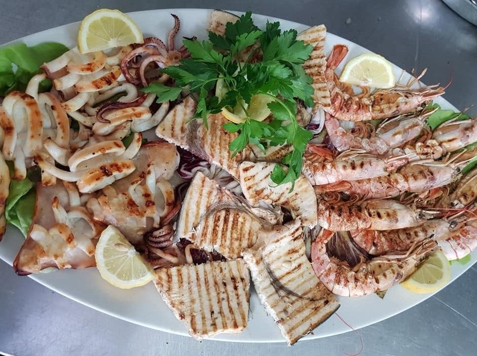 Piatto misto di frutti di mare grigliati con gamberi, pesce e spicchi di limone su un piatto ovale bianco.