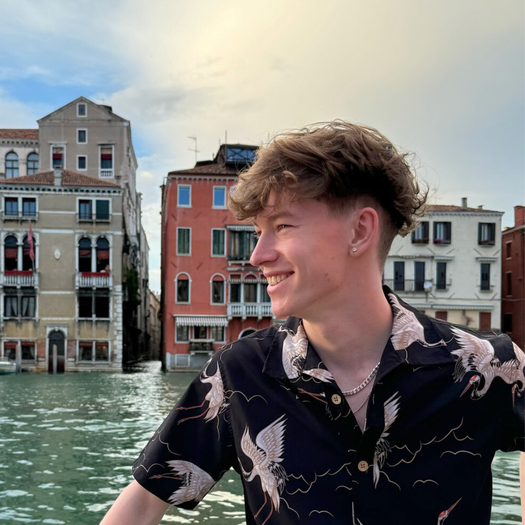 A young man is standing in front of a body of water with buildings in the background