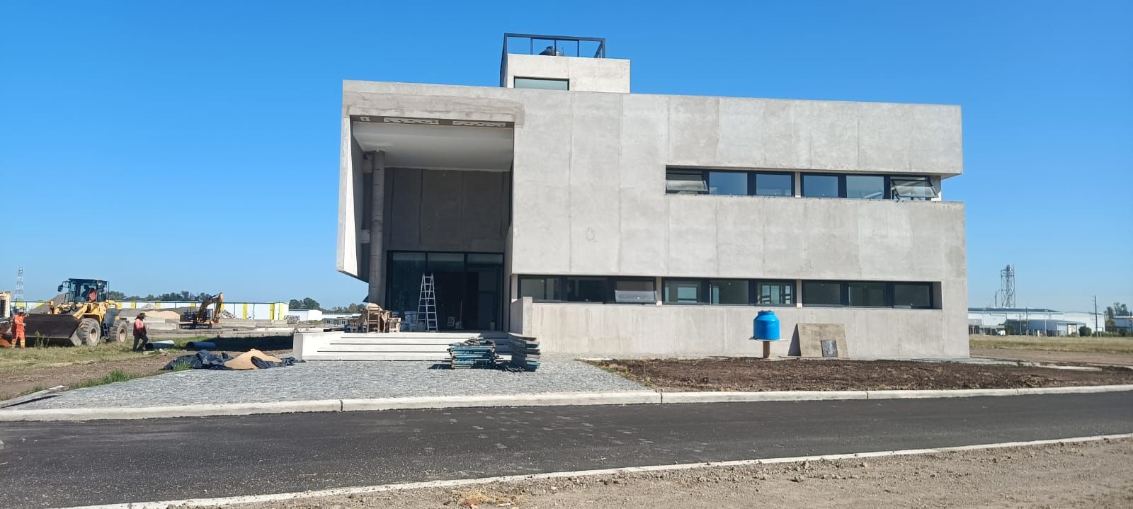 Edificio moderno de hormigón en construcción junto a una carretera asfaltada bajo un cielo azul despejado.