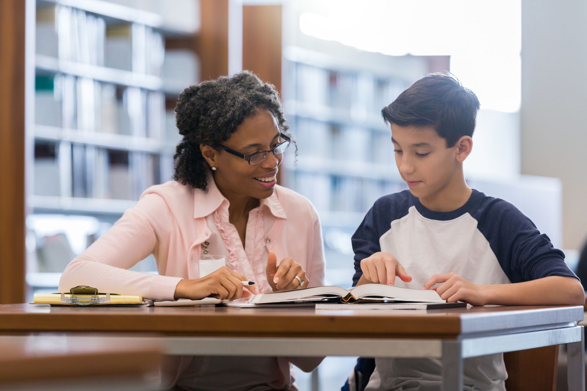 Woman in glasses and boy reading together at a table in a library.