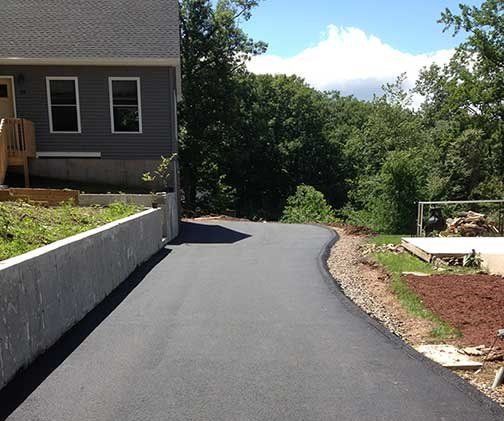 Dark asphalt driveway curves towards a house with gray siding, a sunny, wooded backdrop.