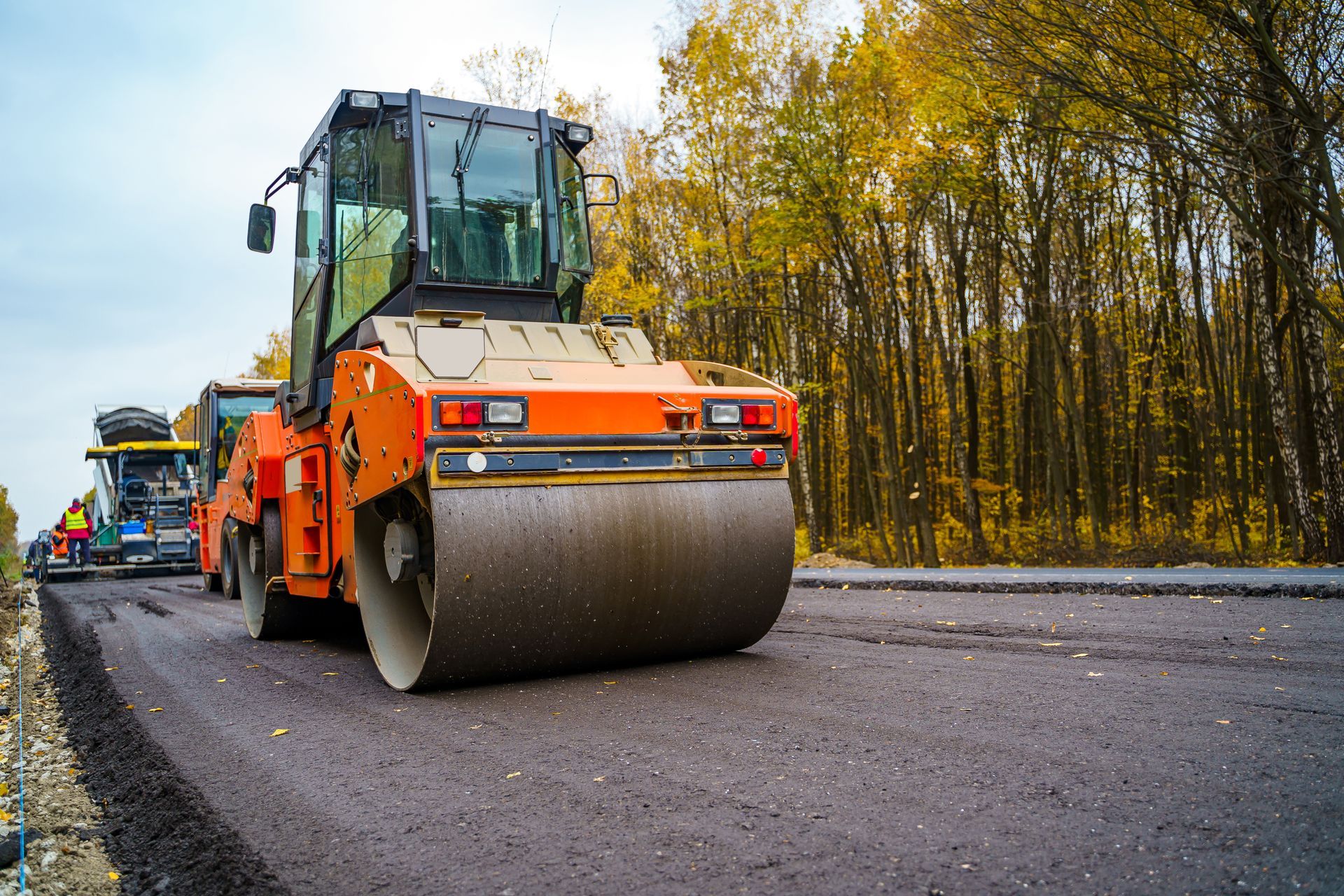 Orange road roller smooths fresh asphalt on a forest lined roadway during paving work. Orange road roller smooths fresh asphalt on a forest lined roadway during paving work.