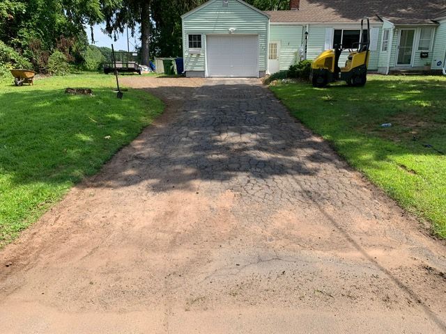 Driveway with gravel and dirt leading to a garage, flanked by grass and a small building.