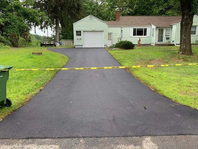 Newly paved asphalt driveway leading to a light green house with a white garage door, grass on either side.