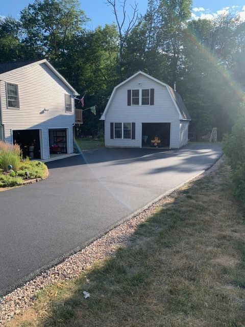 Paved driveway leading to two-story white garage and house, surrounded by grass and trees, bright sunlight.