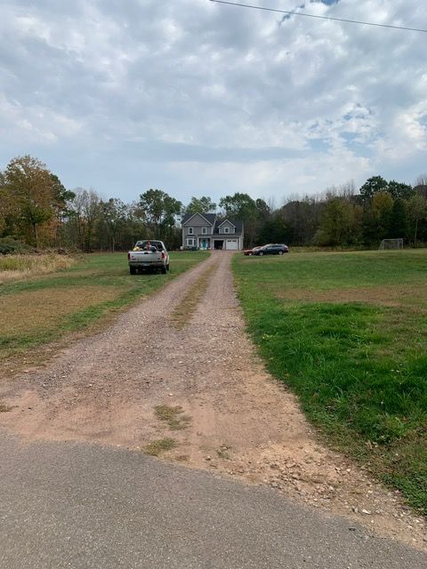 Gravel driveway leads to a gray house with two cars parked in front, surrounded by green grass and trees under a cloudy sky.
