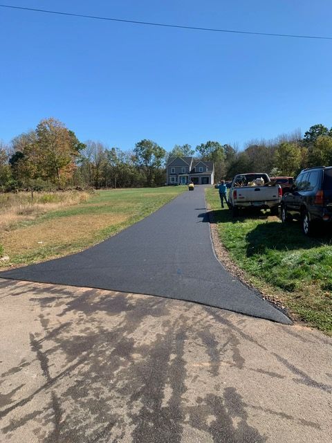 Paved black asphalt driveway leading to a house, with grass on either side and cars parked along the edge.