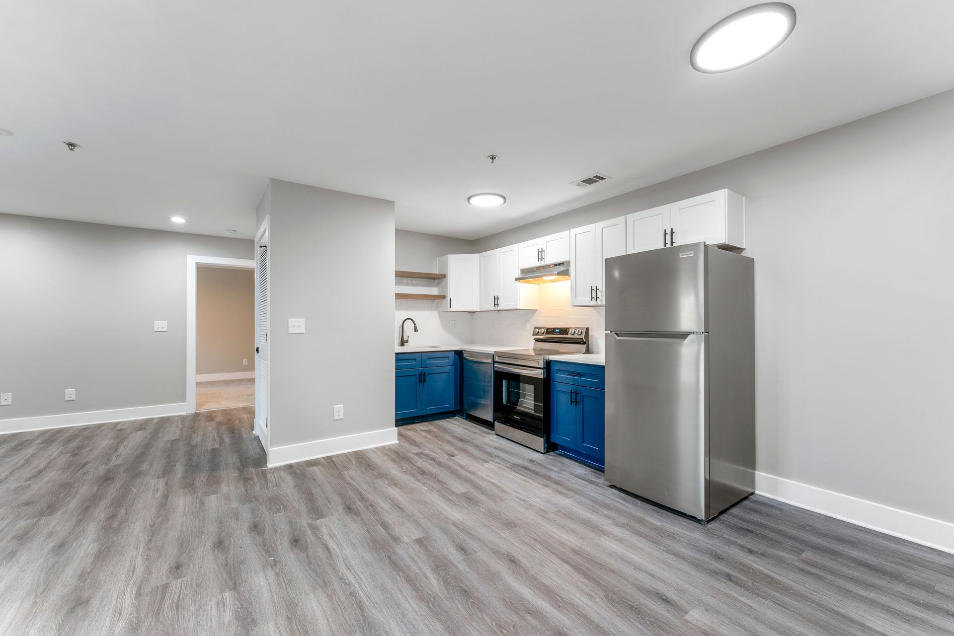 Empty modern kitchen and living area with gray flooring, white cabinets, blue lower cabinets, and a stainless fridge