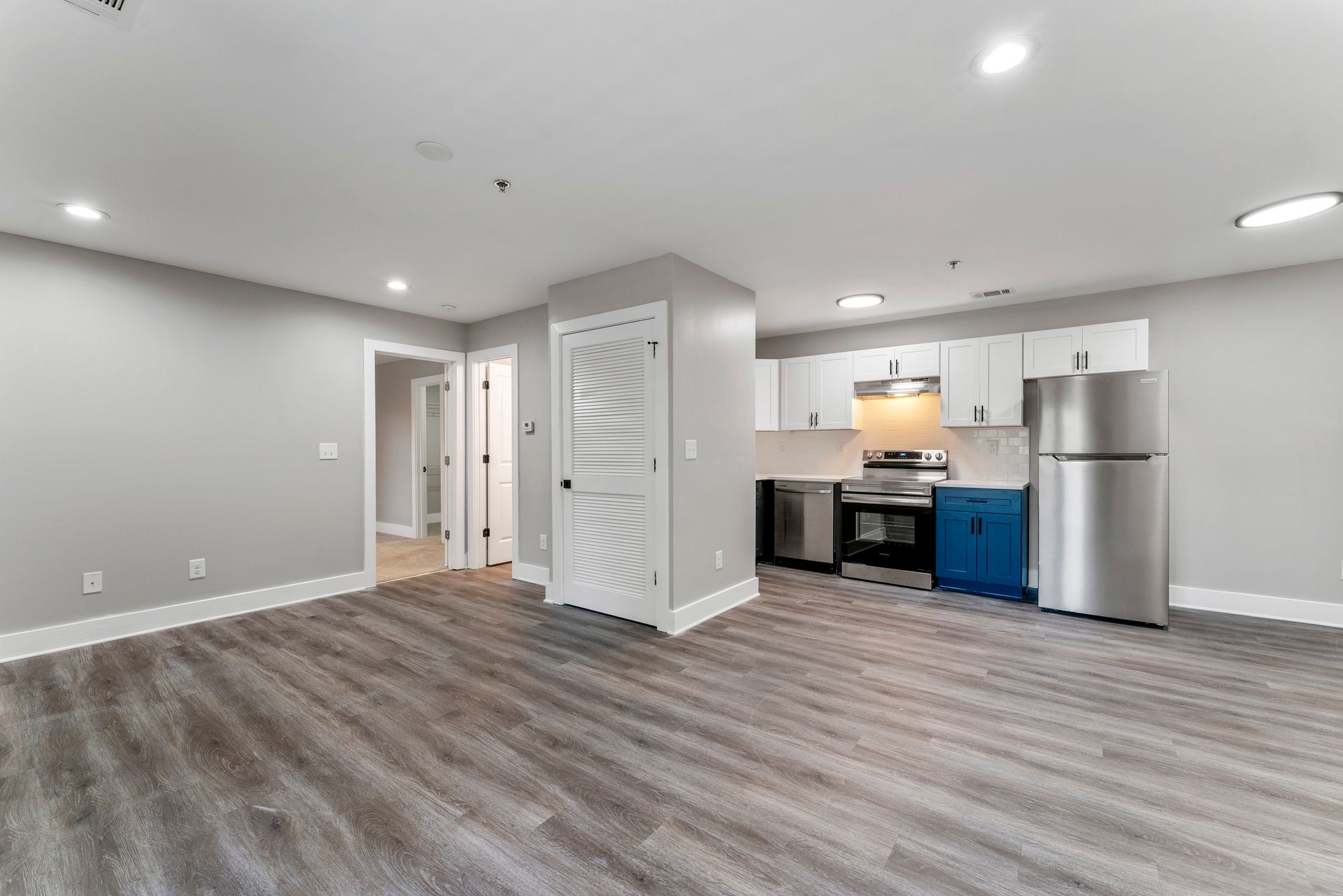 Empty modern basement with gray flooring, white walls, recessed lights, and a small kitchenette with stainless appliances.