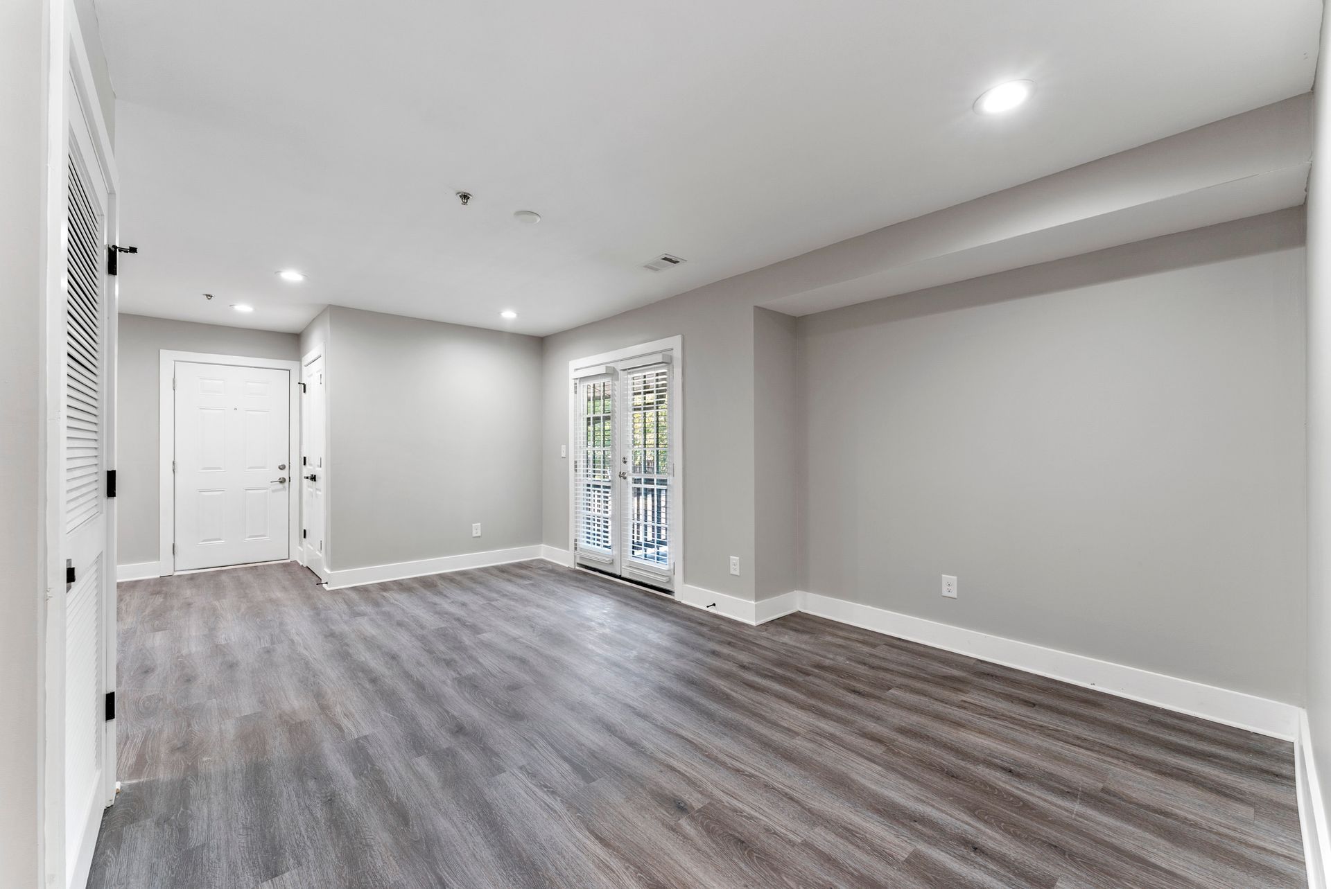 Empty modern living room with gray walls, wood floors, recessed lighting, and a sliding glass door