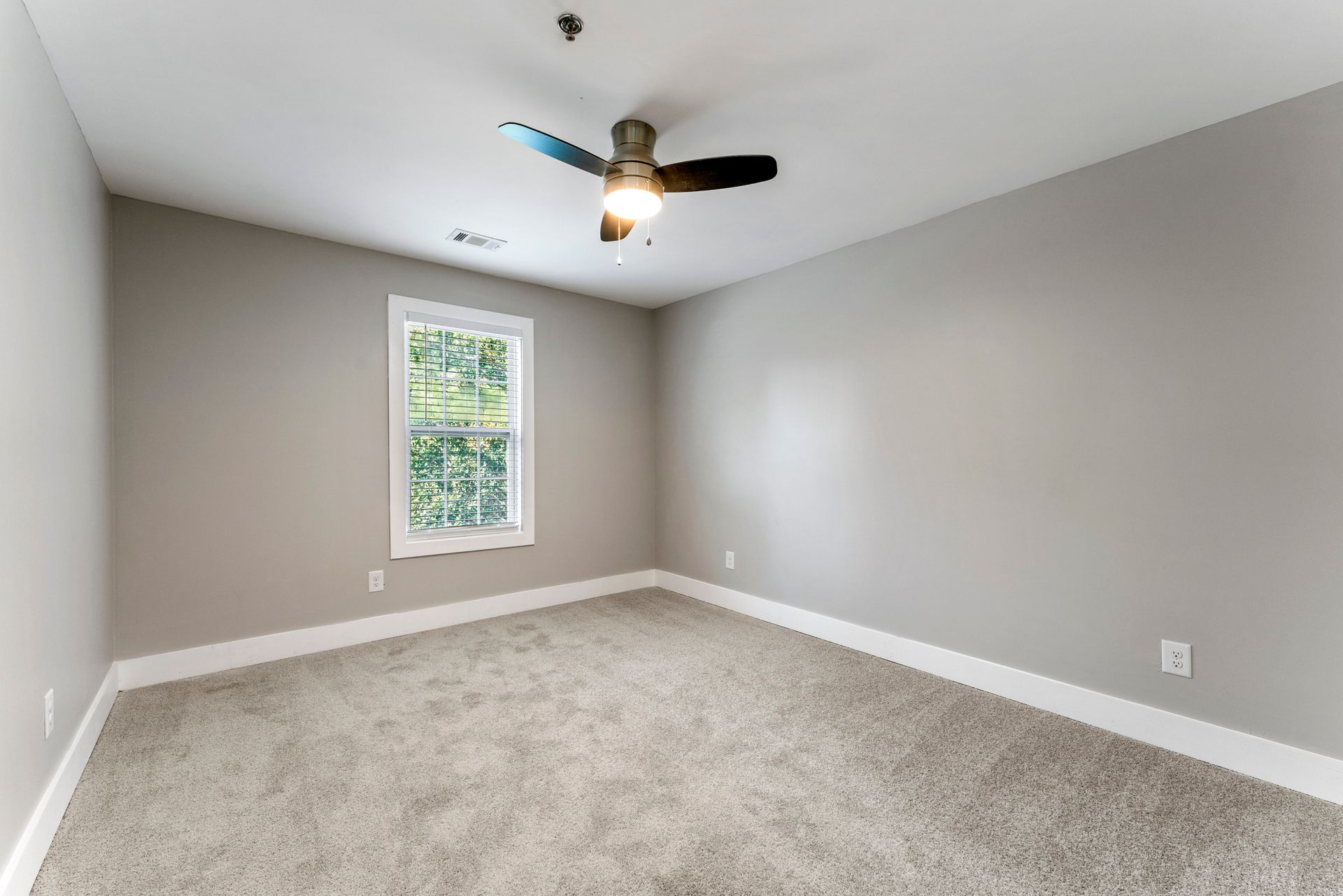 Empty beige bedroom with gray carpet, single window, and ceiling fan.