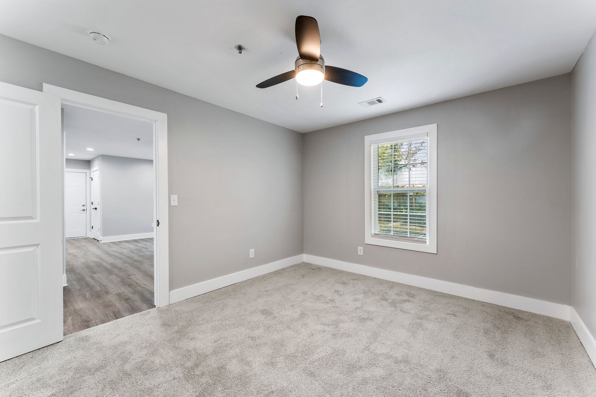 Empty gray bedroom with carpet, ceiling fan, and a window looking into a hallway and another room