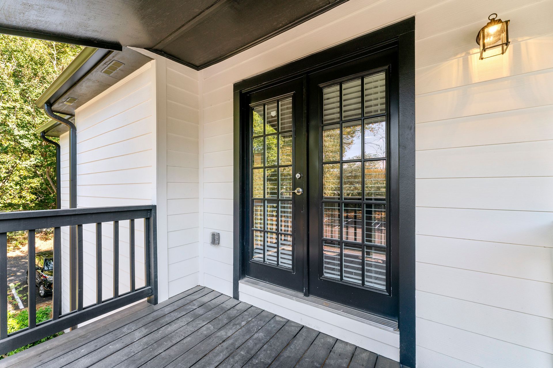 Covered porch with black French doors, white siding, and a dark railing overlooking trees