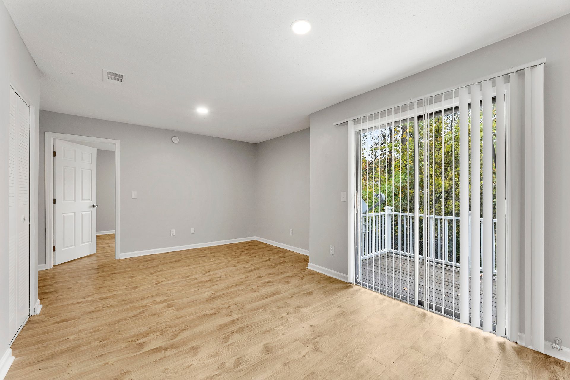 Empty modern room with light wood flooring, gray walls, white trim, and sliding glass doors with vertical blinds