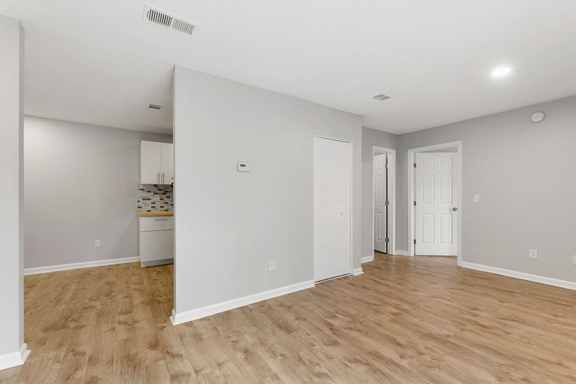 Empty modern living room with wood floors, gray walls, and white trim, opening to a kitchen area