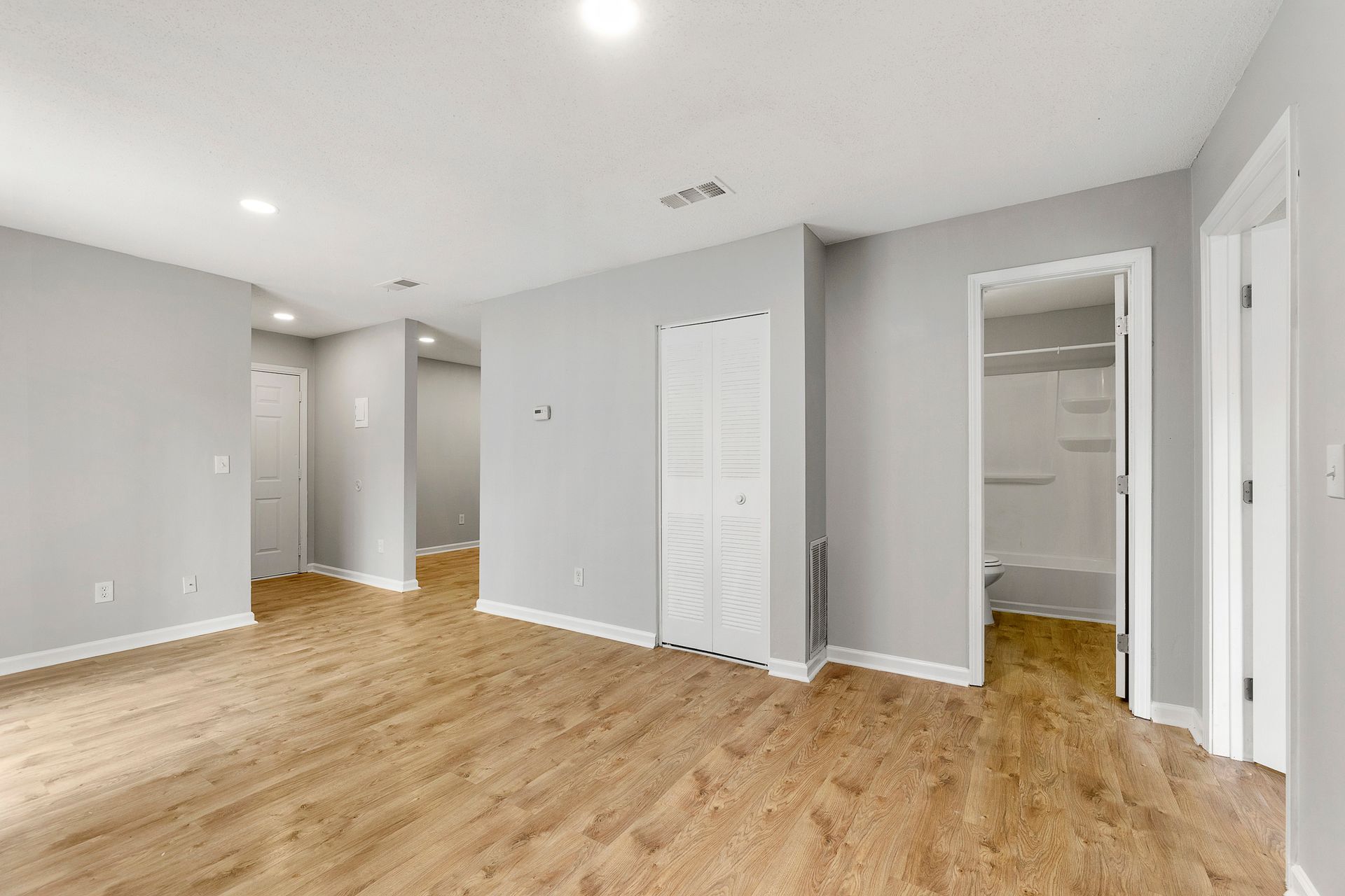 Empty gray bedroom with wood floors, white closet doors, and an open doorway to a bathroom.