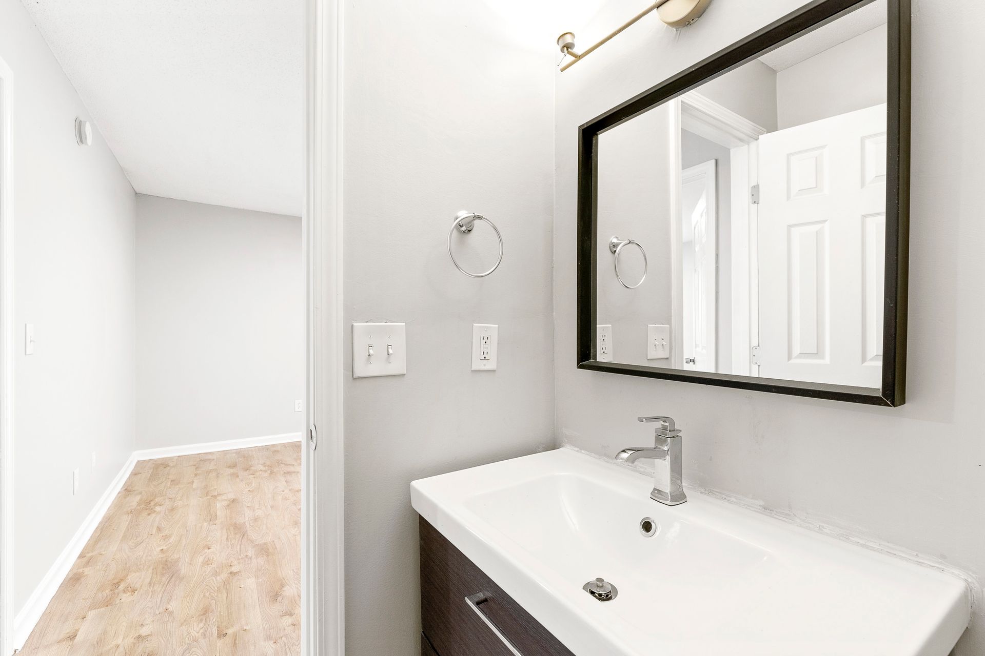 Bright bathroom vanity with mirror and white sink beside a hallway with light wood flooring