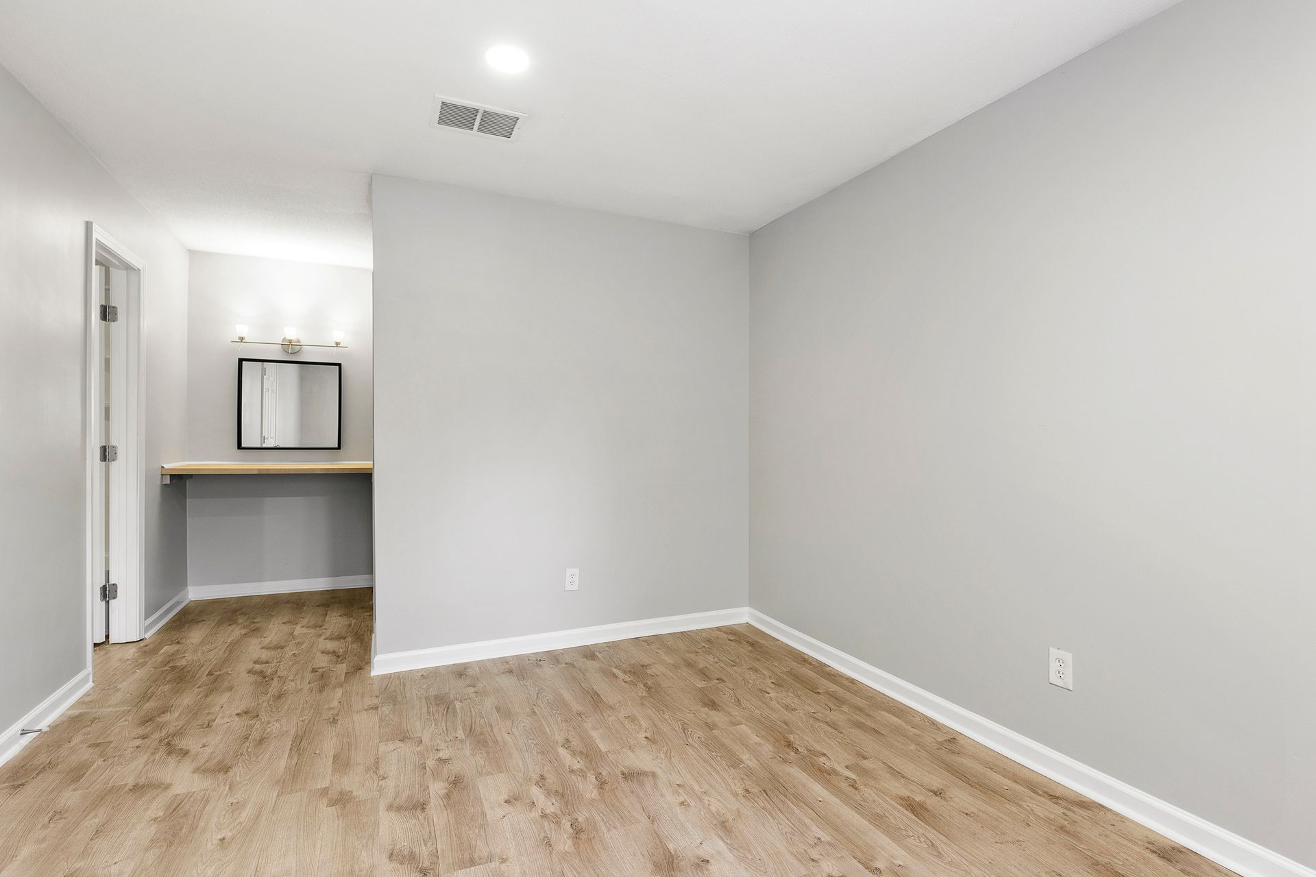 Empty bedroom with light gray walls, wood flooring, and a small vanity area by the hallway