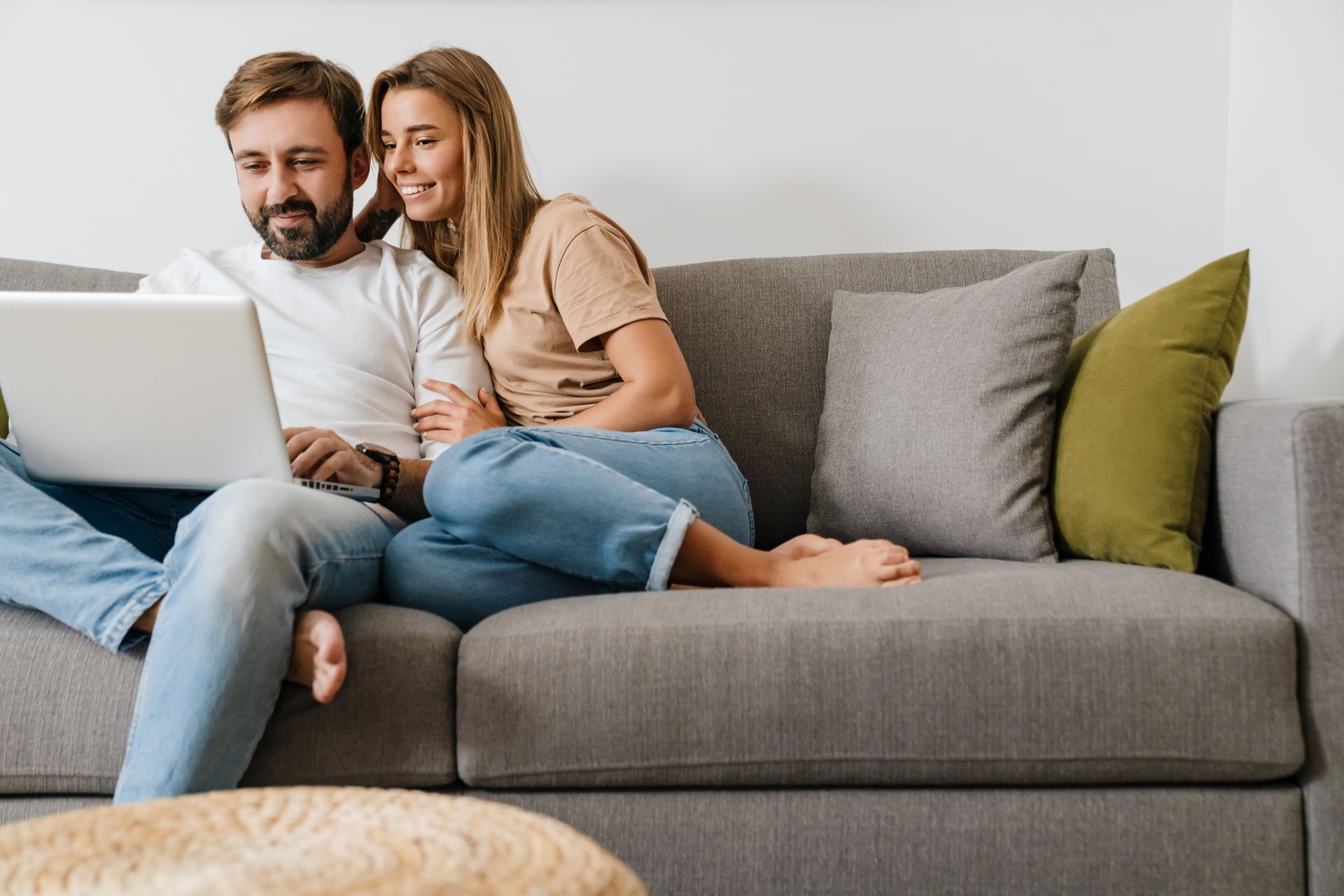 Couple sitting on a gray sofa, looking at a laptop in a bright living room.
