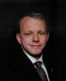 Formal portrait of a man in a dark suit and tie against a black background