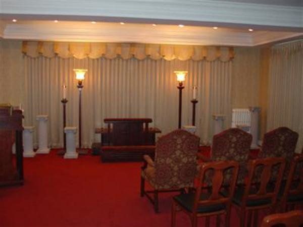 Empty formal room with red carpet, draped curtains, tall lamps, and rows of upholstered chairs facing a front table