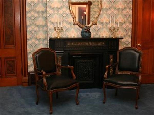 Two dark chairs facing a black fireplace in a wood-paneled room with patterned wallpaper and a blue carpet