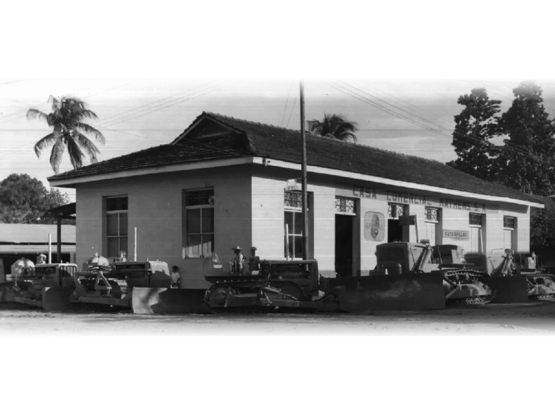 Fotografía en blanco y negro de un pequeño edificio al borde de la carretera con coches antiguos aparcados delante.