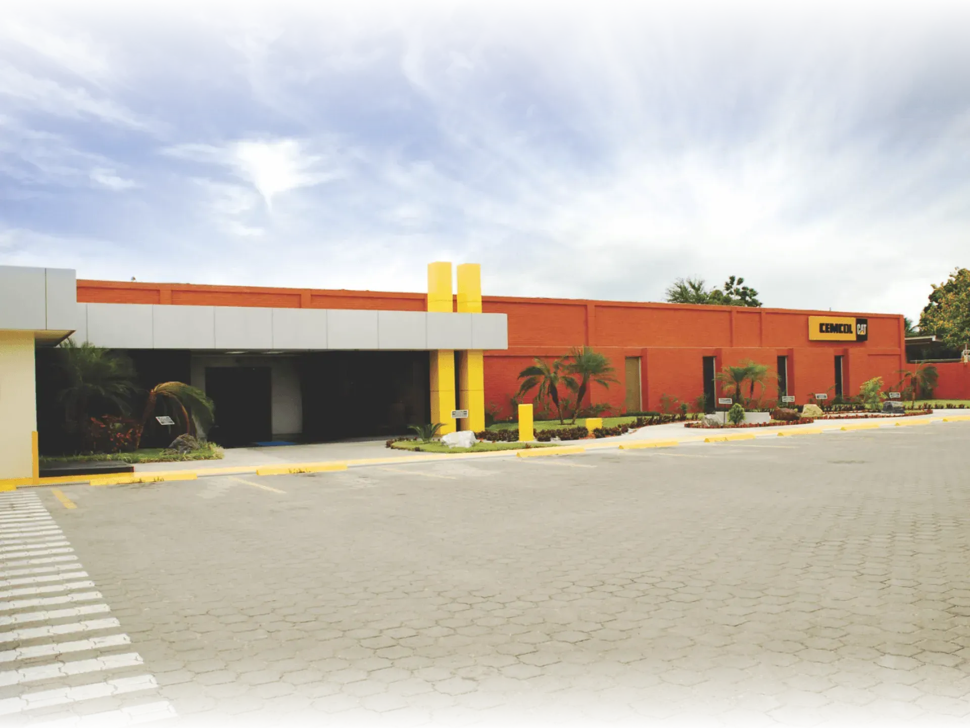 Edificio comercial bajo, de color naranja y blanco, con un estacionamiento bajo un cielo brillante y nublado.