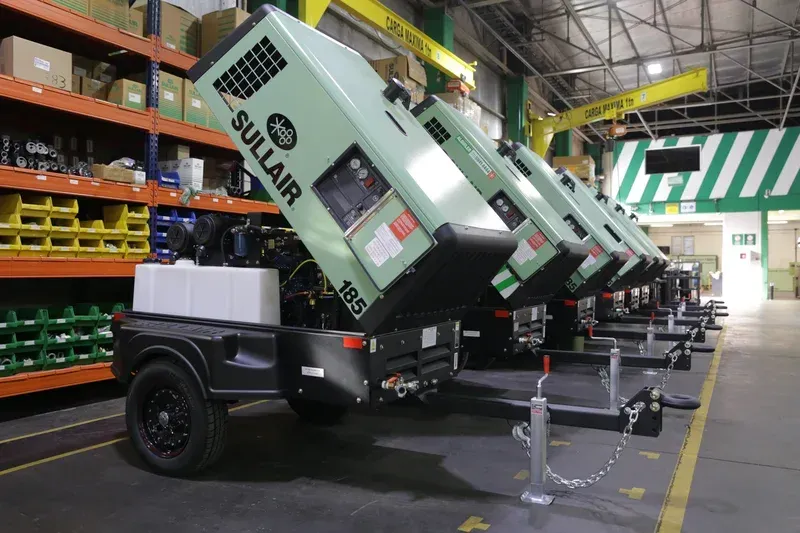 Fila de remolques utilitarios verdes en un almacén, estacionados junto a estanterías y una barandilla de seguridad amarilla.