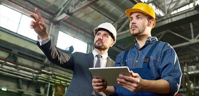 Un hombre con traje y corbata está de pie junto a un trabajador de la construcción en una fábrica mirando una tableta.