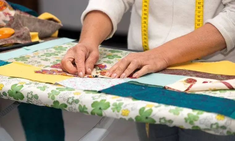 Hands arranging fabric pieces on a quilt with a yellow tape measure in a sewing workspace