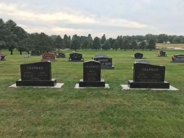 Cemetery lawn with three black gravestones in the foreground and rows of trees and markers behind them