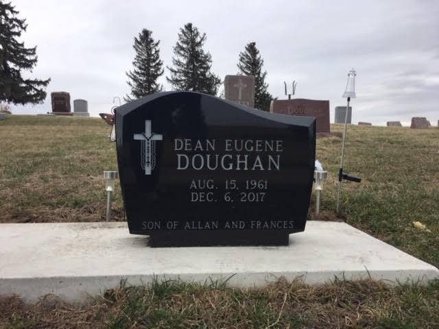 Black gravestone memorial with cross in a cemetery, set on a concrete base with trees in the background